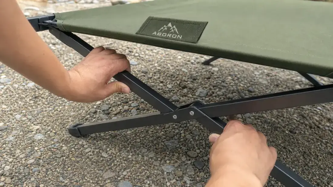 Close-up of hands locking the steel leg frame of a folding camping cot into place at a campsite