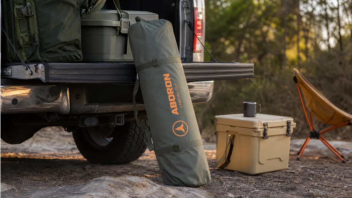Folded camping cot packed into its carry bag leaning against a truck bed at a campsite